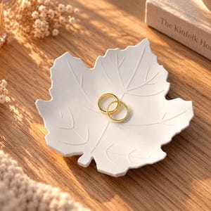 Puede incluir: Un plato decorativo de cerámica blanca con forma de hoja de arce y detalles de nervaduras en relieve. Dos anillos de metal dorado sencillo descansan en el centro del plato, que presenta un acabado mate liso y un borde dentado distintivo.