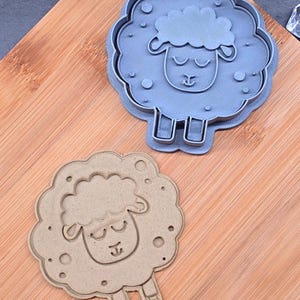 May include: Two sheep-shaped cookie cutters. One is grey and the other is tan. Both feature a sheep face with closed eyes and a fluffy outline. The cutters are on a wooden surface.