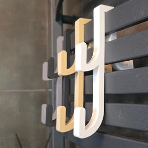 Peut inclure: Un ensemble de trois crochets en forme de J, de couleurs blanc, beige et gris foncé, conçus pour s'accrocher aux barres horizontales d'un radiateur sèche-serviettes. Chaque crochet présente une structure à deux branches, offrant plusieurs points.