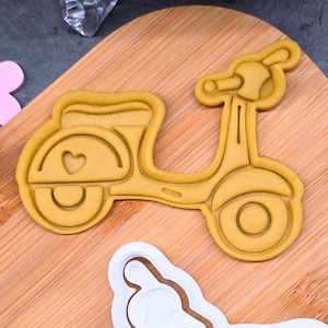 Peut inclure: Un emporte-pièce en forme de scooter jaune avec un détail en forme de cœur. Il est posé sur une planche à découper en bois, avec un autre emporte-pièce blanc en forme de scooter.