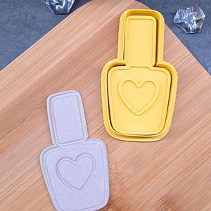 May include: Two nail polish-shaped cookie cutters. One is yellow, the other is light gray. Both cutters have a heart shape in the center. The cutters are on a wooden cutting board.