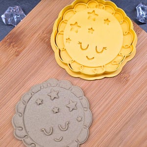 Puede incluir: Dos cortadores de galletas, uno amarillo y otro beige, cada uno con un borde festoneado y un diseño celestial. El cortador amarillo presenta una cara de sol sonriente con estrellas, mientras que el cortador beige tiene el mismo diseño. Ambos están sobre una superficie de madera.
