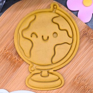 Op de afbeelding: Een gele koekjesvorm in de vorm van een wereldbol met een lachend gezicht. De vorm heeft een gedetailleerde omtrek van de continenten en staat op een standaard. De vorm ligt op een houten snijplank.