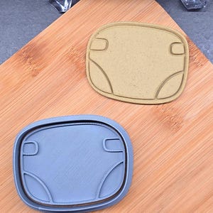 Peut inclure: Deux emporte-pièces, un beige et un bleu, sont posés sur une planche à découper en bois. Les deux emporte-pièces ont la forme d'une couche pour bébé. L'emporte-pièce beige est de couleur unie, tandis que le bleu a une surface texturée.