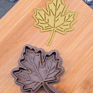 May include: Two maple leaf-shaped cookie cutters. One is a light yellow color, and the other is a dark gray color. The cutters are made of plastic and are designed to cut out the shape of a maple leaf from dough or other materials.