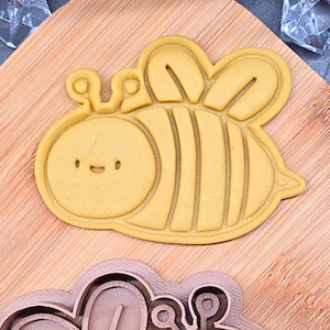 Può includere: Tagliabiscotti a forma di ape gialla con un volto sorridente e un corpo a strisce. Il tagliabiscotti è realizzato in materiale giallo chiaro e poggia su un tagliere di legno. Il design è dettagliato e adatto alla cottura.