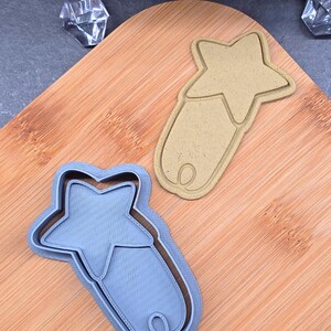 Peut inclure: Deux emporte-pièces en forme d'étoile, un bleu clair et un beige, sont posés sur une planche à découper en bois. Les emporte-pièces ont une étoile en haut et une base arrondie avec une boucle. L'emporte-pièce bleu clair est imprimé en 3D.