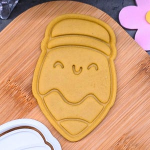 May include: A yellow cookie cutter in the shape of a smiling face with a hat. The cutter is on a wooden cutting board, with a white plastic cutter nearby. The cookie cutter has a happy face design.