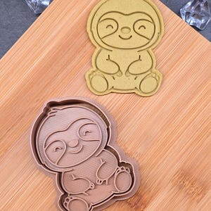May include: A light brown sloth-shaped cookie cutter and a cookie made with the cutter are displayed on a wooden cutting board. The cookie cutter is a light brown color and has a detailed design. The cookie is a similar color and shape.