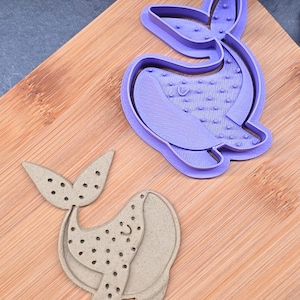 Op de afbeelding: Een paarse walvisvormige koekjessnijder en een beige walvisvormige koek. De koekjessnijder is gemaakt van plastic en heeft een gedetailleerd ontwerp. De beige koek heeft een lachend gezicht en een gestippeld patroon.