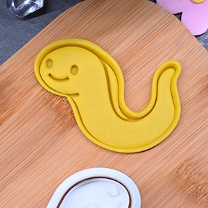 May include: A yellow cookie cutter in the shape of a smiling worm. The cutter is on a wooden cutting board, with a white cookie cutter in the shape of a smiling face below it. A clear, faceted object is in the upper left corner.