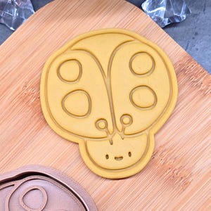 Könnte beinhalten: Ein hellgelber Marienkäfer-förmiger Ausstecher mit einem lächelnden Gesicht und detaillierten Flügelmustern. Der Ausstecher liegt auf einem Holzschneidebrett, mit einem zweiten, ähnlichen Ausstecher darunter.