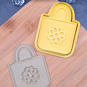 Può includere: Due formine per biscotti a forma di borsa con un motivo floreale. Uno è giallo, l'altro è grigio chiaro. Le formine sono su un tagliere di legno.