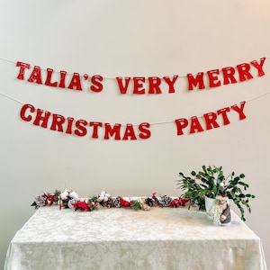 May include: A festive Christmas party scene with a red banner that reads "TALIA'S VERY MERRY CHRISTMAS PARTY." A table is set with a white tablecloth, decorated with a garland of pinecones, cotton, and red accents. A Santa Claus figurine and greenery complete the holiday decor.