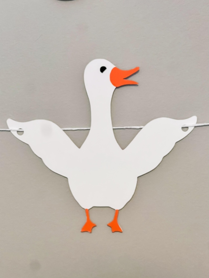 May include: White paper cutout of a goose with orange beak and feet. The goose is hanging from a string.