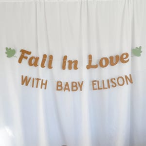 May include: A banner with the words "Fall In Love" in brown script above "With Baby Ellison" in block letters. The banner is strung on a white string with green leaf accents, set against a white backdrop.