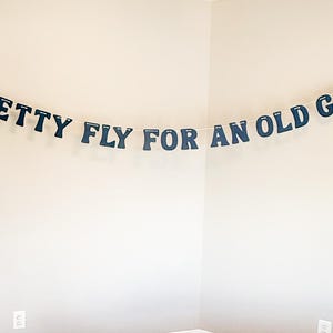 May include: A navy blue banner strung across a wall. The banner reads "PRETTY FLY FOR AN OLD GUY" in uppercase letters. The letters are attached to a white string and are hanging in a room with light gray walls.