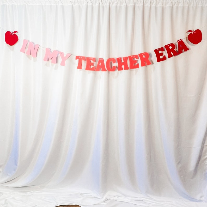 May include: A banner with the text "IN MY TEACHER ERA" in red and pink letters, with red apple cutouts on each end. The banner is strung across a white curtain backdrop.