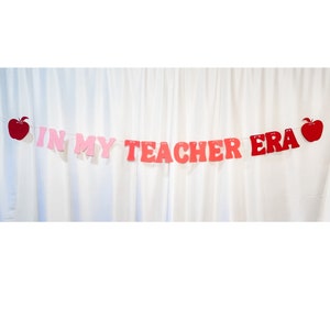 May include: A banner with the text "IN MY TEACHER ERA" in pink and red letters, strung between two red apple cutouts. The banner is displayed against a white curtain backdrop.