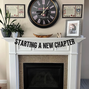 May include: A white fireplace mantel with a black banner that reads "STARTING A NEW CHAPTER". The banner is hanging above the fireplace. There is a clock, two framed pictures, and a wooden bowl on the mantel.