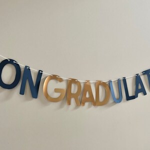 K&ouml;nnte beinhalten: Ein marineblaues und goldenes Banner mit dem Wort "CONGRATULATIONS" auf einer wei&szlig;en Schnur.