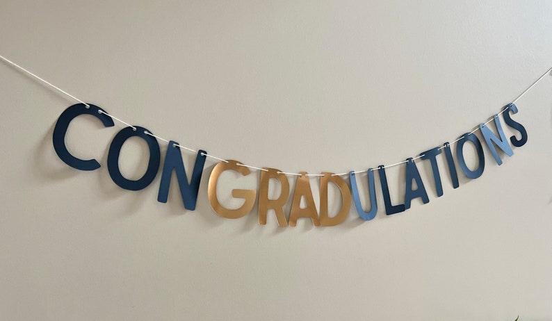 K&ouml;nnte beinhalten: Ein marineblaues und goldenes Banner mit dem Wort "CONGRATULATIONS" auf einer wei&szlig;en Schnur.
