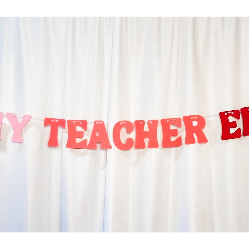 May include: A banner with the words "MY TEACHER" in red letters and "ER" in red letters, strung on a white string. The letters are in a bold, rounded font. The background is a white curtain.