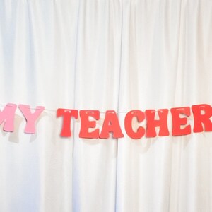 May include: A banner with the phrase "IN MY TEACHER ERA" in red and pink letters, strung between two red apple cutouts. The banner is displayed against a white curtain backdrop, suitable for classroom or teacher-themed decor.