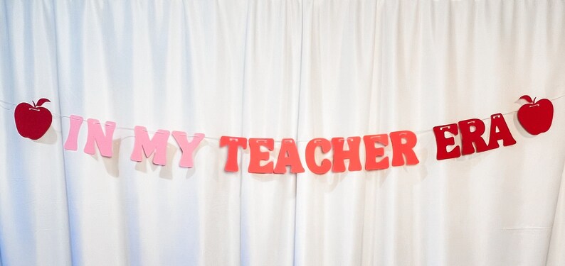 May include: A banner with the phrase "IN MY TEACHER ERA" in red and pink letters, strung between two red apple cutouts. The banner is displayed against a white curtain backdrop, suitable for classroom or teacher-themed decor.