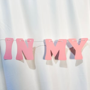 May include: A pink banner with the words "IN MY" strung across a white string. The letters are in a bold, rounded font and are a soft, pastel pink color. The banner is hanging against a white backdrop.