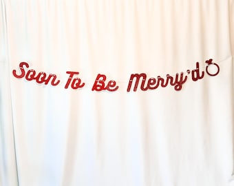 May include: Red glitter banner with the words "Soon To Be Merry'd" in a cursive font, with a ring graphic at the end. The banner is strung on a thin white string and hangs against a white backdrop.
