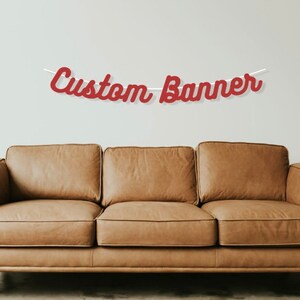 K&ouml;nnte beinhalten: Ein rotes individuelles Banner mit dem Text "Custom Banner" h&auml;ngt &uuml;ber einem braunen Ledersofa.