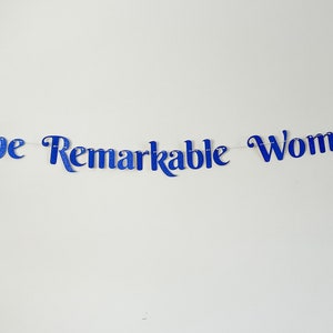 Puede incluir: Pancarta azul con purpurina con el texto "One Remarkable Woman" colgada en una pared blanca.