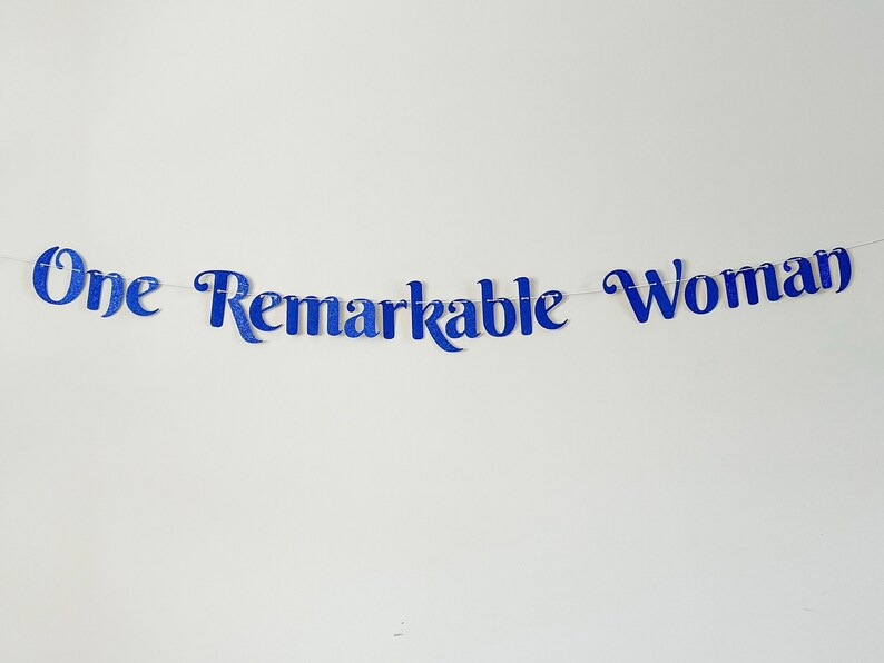 Puede incluir: Pancarta azul con purpurina con el texto "One Remarkable Woman" colgada en una pared blanca.