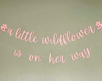 May include: A light pink banner featuring the text "a little wildflower is on her way" in a cursive script font. The banner is divided into two lines and includes two small flower cutouts at the ends of the top line, all strung on thin wire.