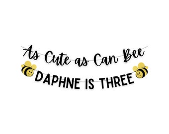 May include: A black banner with the text "As Cute as Can Bee" and "Daphne is Three". The banner is decorated with two yellow and black bee illustrations. The banner is strung on a thin silver string.