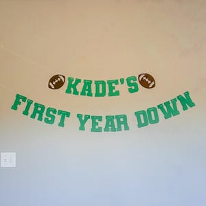 May include: A green banner with the text "KADE'S FIRST YEAR DOWN" and two brown footballs. The banner is strung across a wall, perfect for a sports-themed birthday party or celebration.