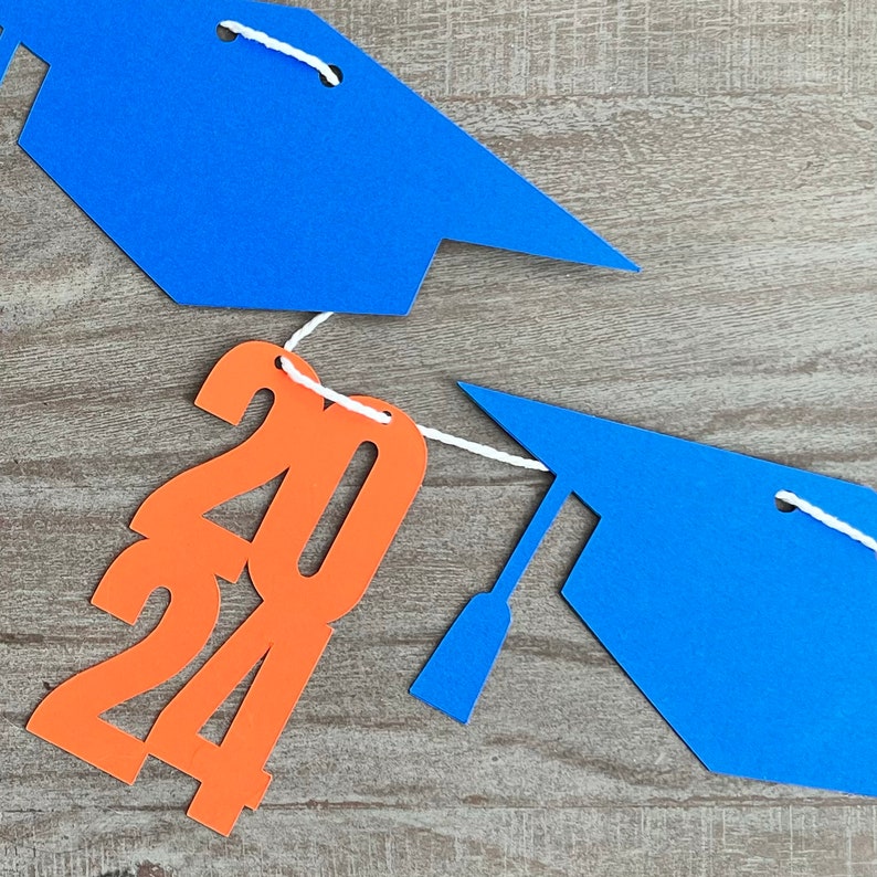 K&ouml;nnte beinhalten: Orangefarbene Papierschnitte der Zahlen 2024 mit blauen Abschlusskappen auf beiden Seiten, verbunden durch eine wei&szlig;e Schnur.