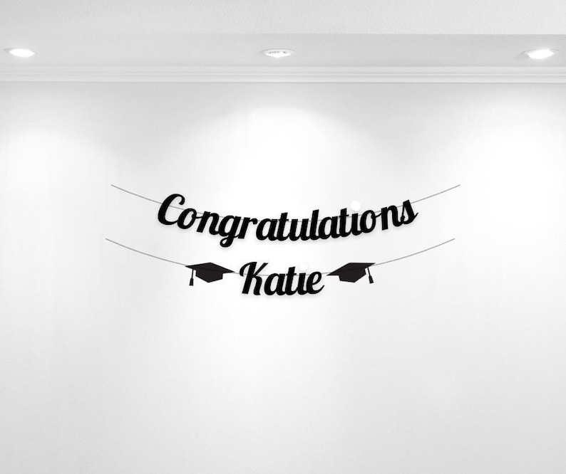 K&ouml;nnte beinhalten: Schwarzer Abschlussbanner mit dem Text "Congratulations Katie" und zwei Abschlusskappen auf beiden Seiten des Namens.
