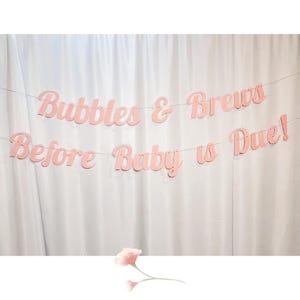 May include: Pink paper banner with the text "Bubbles & Brews Before Baby is Due!" hanging on a white background.