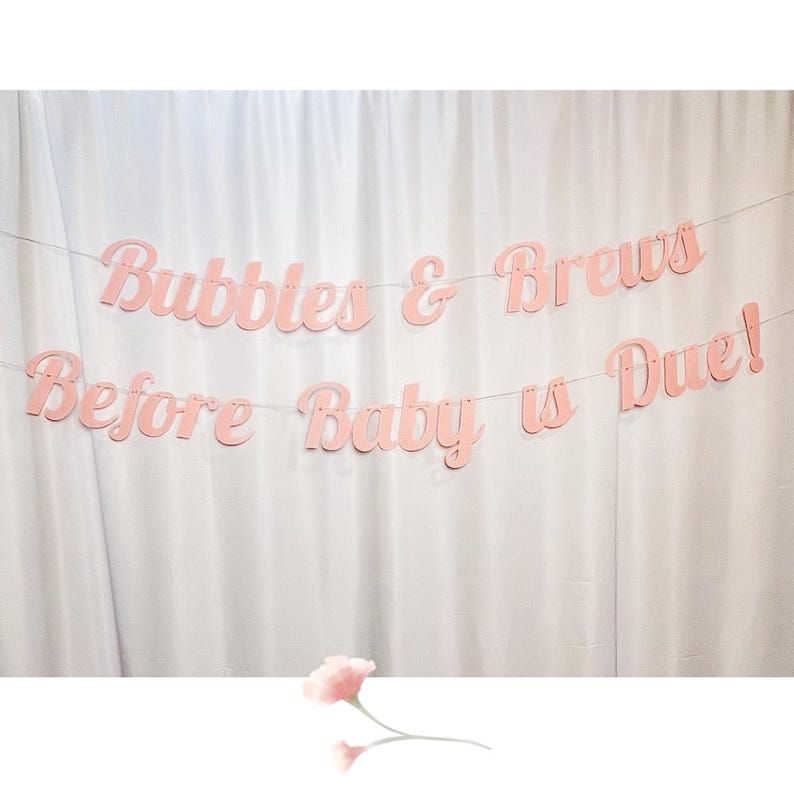 May include: Pink paper banner with the text "Bubbles & Brews Before Baby is Due!" hanging on a white background.