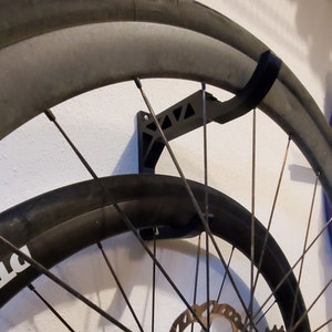May include: A black bike wheel storage rack mounted on a white wall. The rack is made of plastic and has a hook to hold the wheel. The wheel is black with silver spokes and a black tyre.