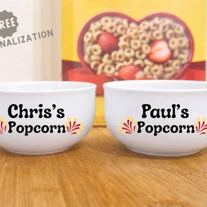 May include: Two white ceramic bowls with the text "Chris's Popcorn" and "Paul's Popcorn" in black, with a decorative yellow, red, and white design. A cereal box is in the background.