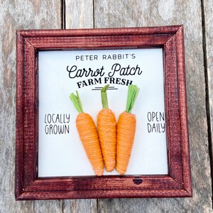 May include: A framed sign with the text "Peter Rabbit's Carrot Patch Farm Fresh" and the words "Locally Grown" and "Open Daily". Three orange yarn carrots are displayed on the sign.