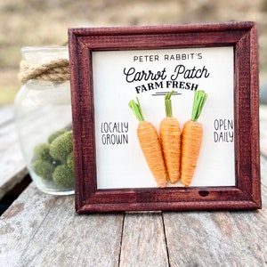 May include: A wooden framed sign with the text "Peter Rabbit's Carrot Patch Farm Fresh" and "Locally Grown" and "Open Daily". Three orange carrots with green tops are displayed on the sign.