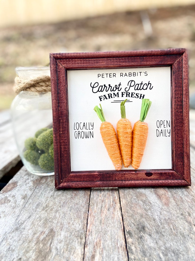May include: A wooden framed sign with the text "Peter Rabbit's Carrot Patch Farm Fresh" and "Locally Grown" and "Open Daily". Three orange carrots with green tops are displayed on the sign.