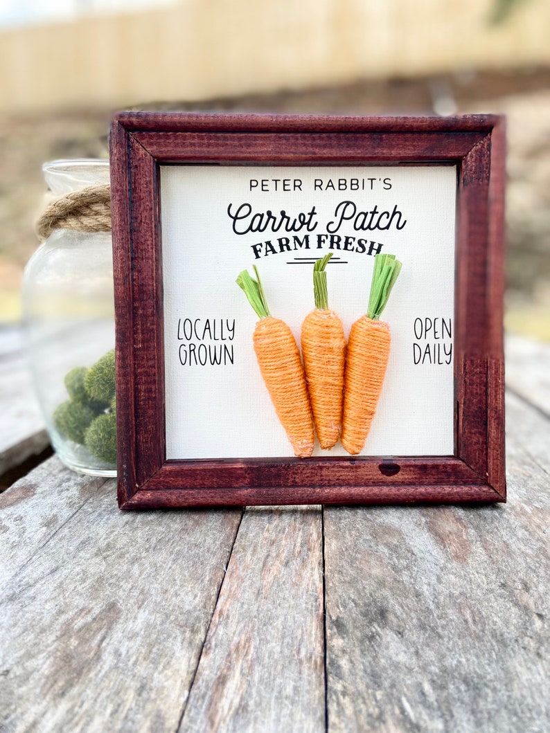 May include: A framed sign with the text "Peter Rabbit's Carrot Patch Farm Fresh" and the words "Locally Grown" and "Open Daily". Three yarn carrots are displayed on the sign.