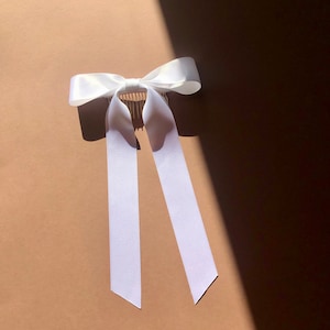 Könnte beinhalten: Weißes Satinband-Haarband mit einem Kamm. Die Schleife ist in einem einfachen Knoten gebunden und die Bänder sind lang und fließend.