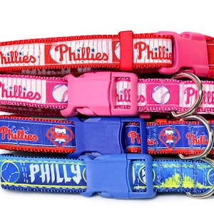 May include: A stack of dog collars in red, pink, blue, and yellow. The collars feature the word "Phillies" and baseball-themed graphics. Each collar has a plastic buckle and a metal D-ring.