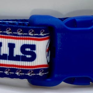 May include: A blue and white dog collar featuring the Buffalo Bills logo and team name. The collar has a blue plastic buckle and a silver metal D-ring. The design includes the team's helmet and charging buffalo.